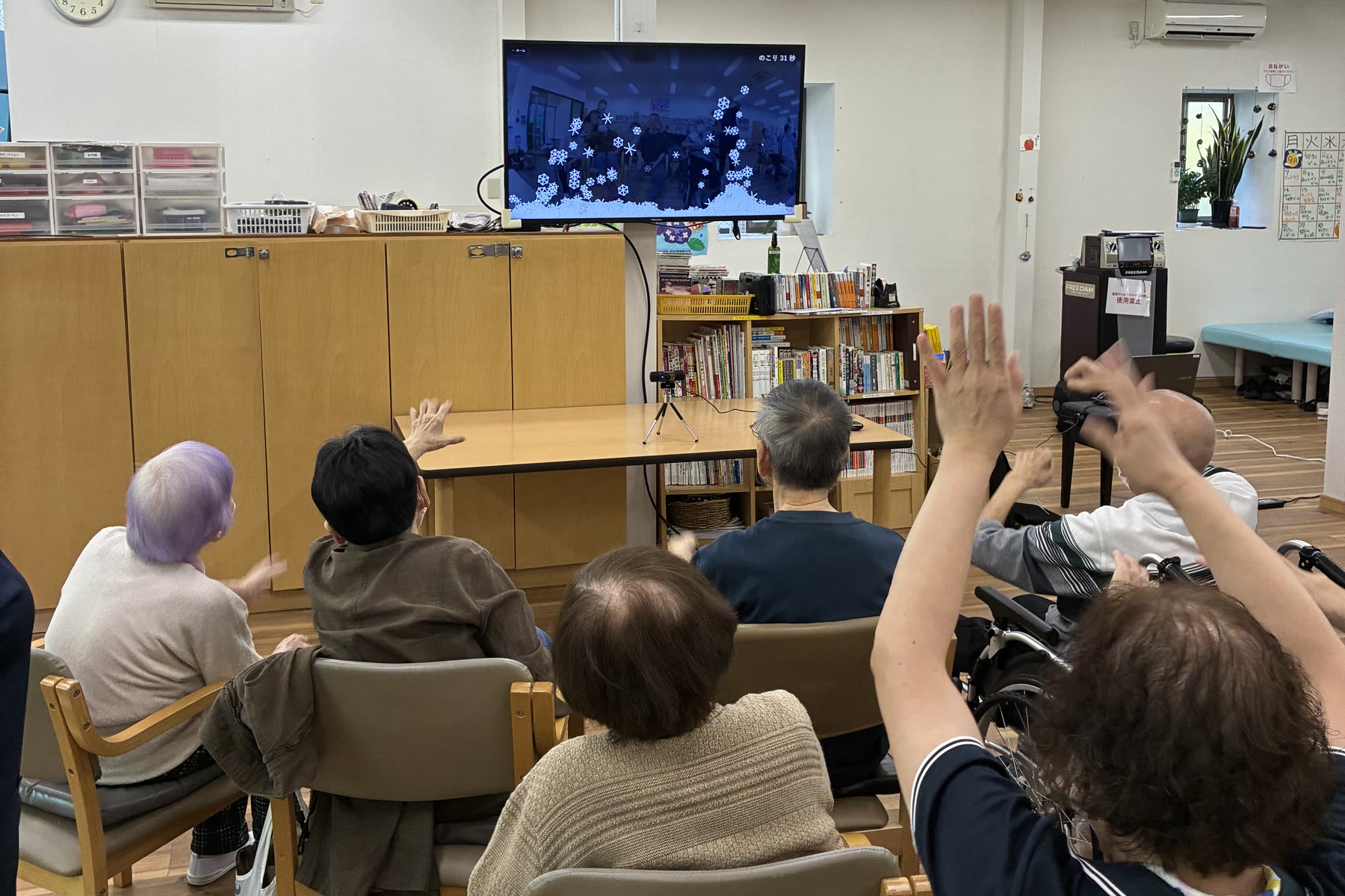 大樹デイサービス流山での体験会。椅子に座った利用者の方々が、テレビの画面に向かって手を上げている後ろ姿